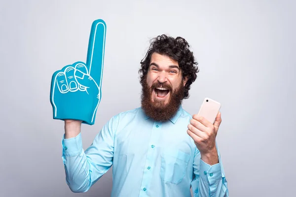 Photo de l'homme barbu étonné en chemise bleue tenant le téléphone et le gant de mousse de ventilateur — Photo