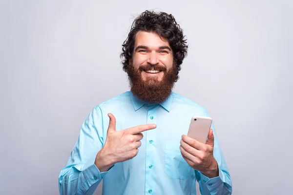 Foto de un joven barbudo hipster sonriente apuntando a un teléfono inteligente sobre una pared blanca — Foto de Stock