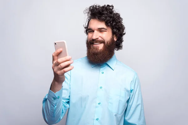 Joven y alegre hombre hipster barbudo con camisa azul usando su teléfono inteligente y sonriendo — Foto de Stock