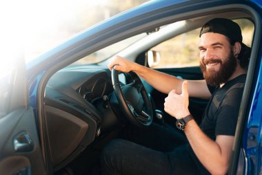 Yakışıklı sürücünün fotoğrafı. Başparmağını gösteriyor, arabada oturuyor ve kameraya bakıyor.