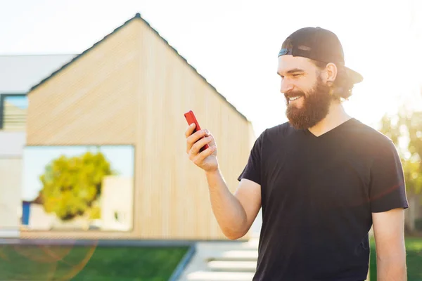 Portrait of handsome man with beard sitting outdoor near house and using smartphone — Stock Photo, Image