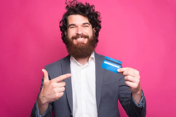 Foto de un hombre feliz con barba usando traje y apuntando a la tarjeta de crédito azul — Foto de Stock