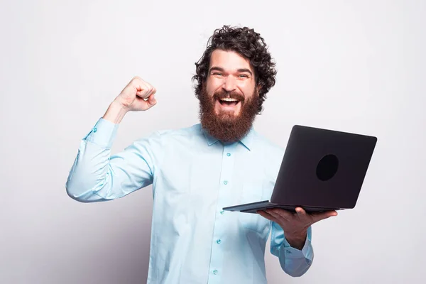 Homem barbudo camisa azul segurando laptop e celebrando a vitória sobre a parede branca — Fotografia de Stock