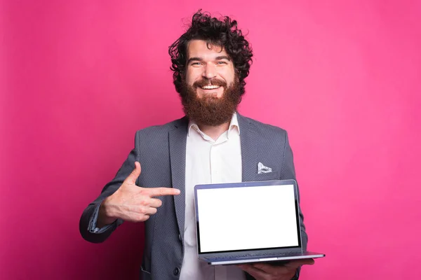 Happy bearded man in suit pointing at blank white screen on laptop and smiling