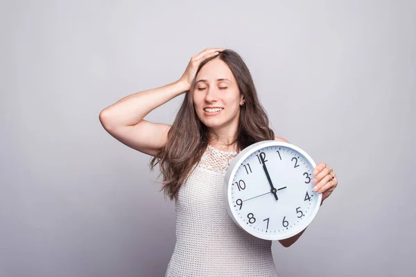 Mujer joven bonita sonríe y sostiene un reloj redondo blanco cerca de una pared gris —  Fotos de Stock