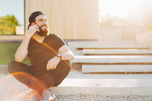 A young bearded man talking to a phone and smiling near his house — Stock Photo, Image