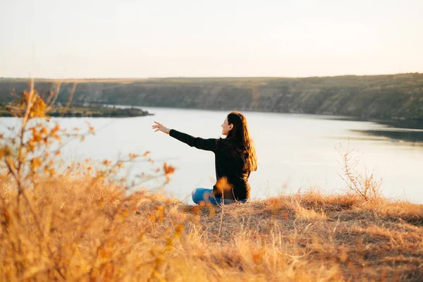 Фото женщины, сидящей на холме возле реки во время заката и указывающей куда-то — стоковое фото