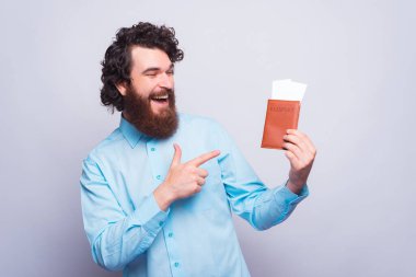 Elinde pasaport olan neşeli bir gencin fotoğrafı. Biletleri gri bir duvarın yanında.