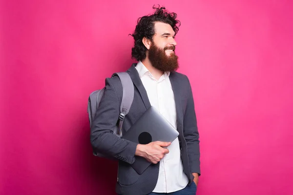 Junger fröhlicher Mann mit Rucksack und neuem Computer lächelt und schaut weg mit einer rosafarbenen Wand hinter sich — Stockfoto