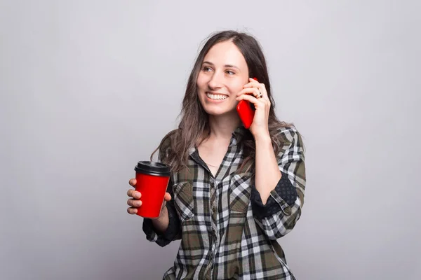 Una mujer hablando con su teléfono sonriendo y sosteniendo una taza caliente para beber cerca de una pared gris — Foto de Stock