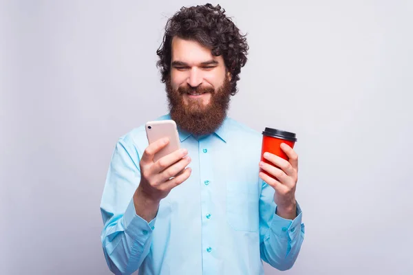 Um homem está olhando e sorrindo para seu telefone e está segurando uma xícara com bebida quente — Fotografia de Stock