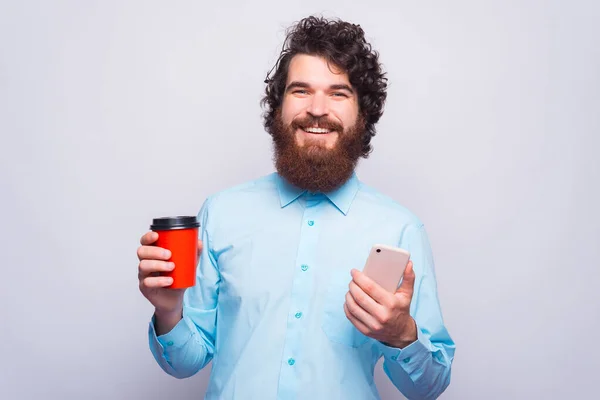 Jovem alegre sorrindo e olhando para a câmera está segurando uma xícara com bebida quente e um telefone — Fotografia de Stock