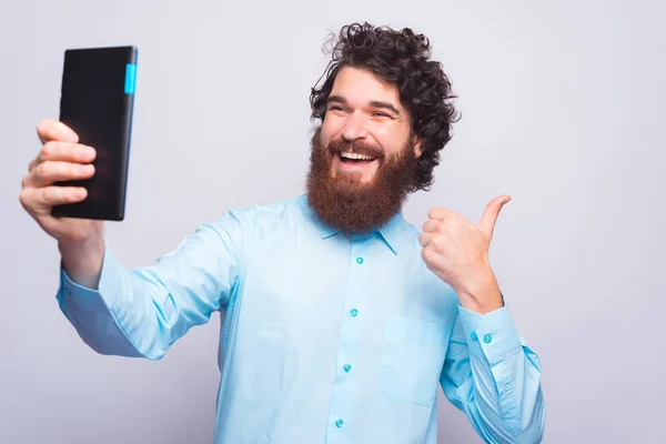 Jovem alegre está segurando um polegar para cima e tomando uma selfie com almofada perto de uma parede cinza — Fotografia de Stock