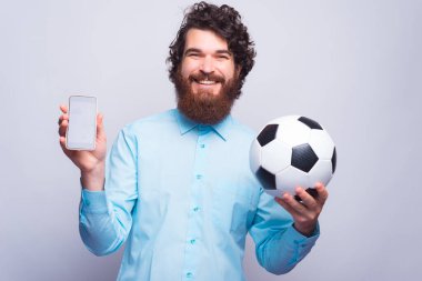 Şu muhteşem uygulamaya bak, sakallı, neşeli, sıradan bir adam ekrandaki telefonu gösteriyor ve elinde futbol topu tutuyor.