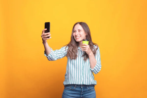 Nice young woman taking a selfie and holding a cup with hot drink near a yellow wall