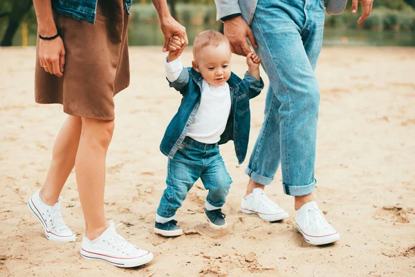 Baby holding his mothers and fathers hands trying to walk in the park — Stock Photo, Image