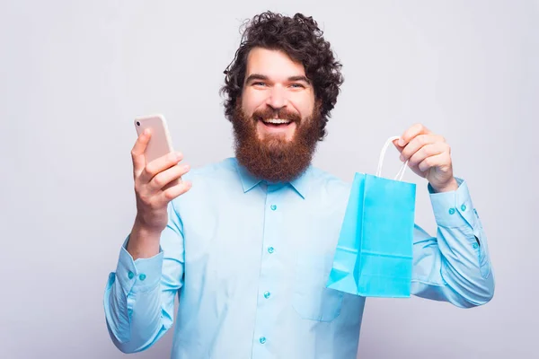 Compras on-line é grande, homem barbudo jovem em camisa azul usando smartphone e segurando saco de compras azul — Fotografia de Stock