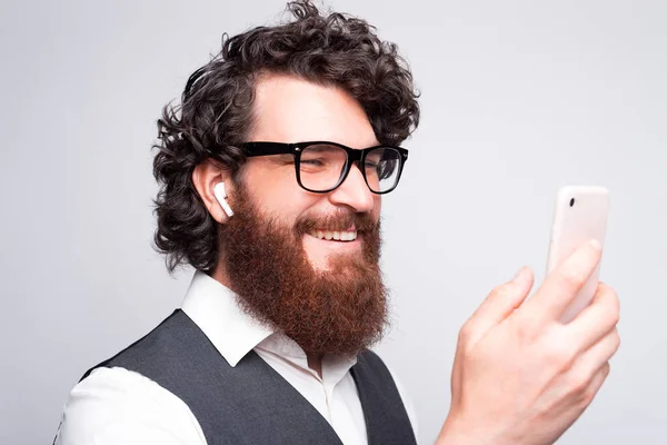 Close up portrait of young bearded hipster man using smartphone and earpods over white wall — Stock Photo, Image