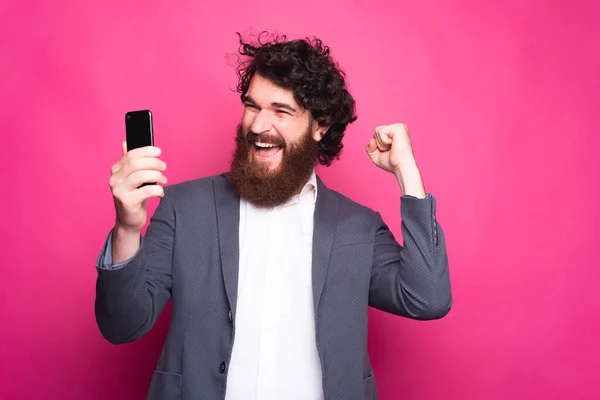 Uma foto de um jovem homem barbudo alegre sendo animado olhando em seu telefone com uma mão no ar perto de uma parede rosa — Fotografia de Stock