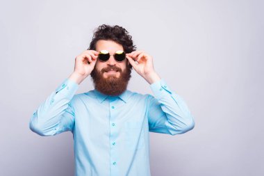 Sakallı genç adam iki eliyle güneş gözlüğünü beyaz bir duvarın yanında tutuyor.