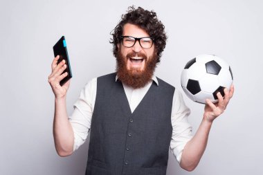 Sakallı ve gözlüklü genç bir adam tablet tutuyor ve bir futbol topu çığlık çığlığa kameraya bakıyor beyaz bir duvarın yanında heyecanlanıyor