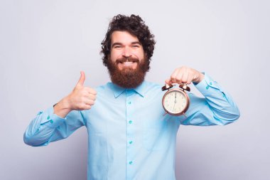 Sakallı, neşeli genç adam başparmağını kaldırıyor ve beyaz bir duvarın yanında bir saat tutuyor.