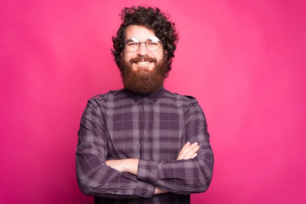 En trevlig ung man med skägg står med armarna korsade nära en rosa vägg med glasögon på — Stockfoto
