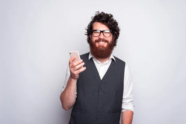 Bearded man with glasses is smiling at the camera and holding a phone near a white wall — Stock Photo, Image