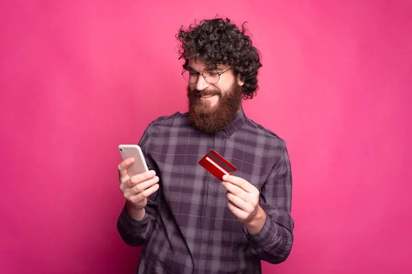戴眼镜的快乐男人笑着在网上买东西，手里拿着手机和信用卡站在粉红的墙上 — 图库照片
