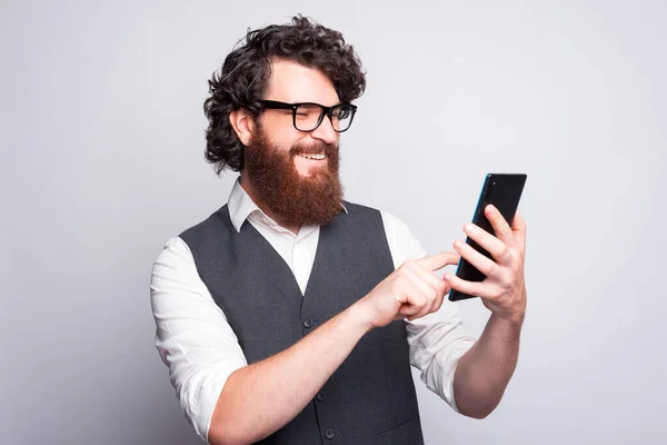 Bearded young man with glasses is smiling and typing in a tablet near a white wall — Stock Photo, Image