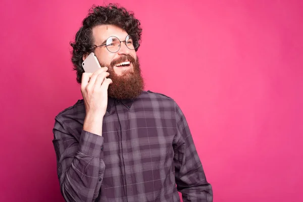 Egy vidám, szakállas fiatalember beszél a telefonjához, és szemüvegben nevet a rózsaszín fal mellett. — Stock Fotó