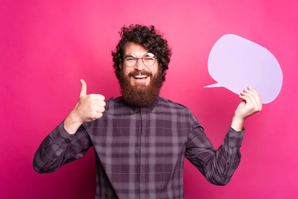 Der bärtige, fröhliche Mann zeigt lächelnd den Daumen nach oben und eine Sprechblase mit seiner Brille an einer rosafarbenen Wand — Stockfoto