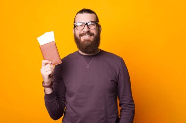 Sakallı adam kameraya gülümsüyor ve elinde içinde iki bilet olan bir pasaport tutuyor.