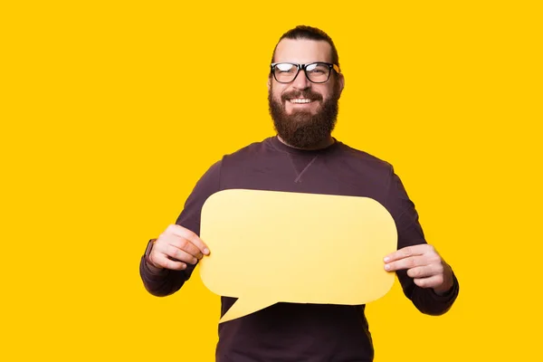 Jeune homme barbu regardant et souriant à la caméra tient une bulle de parole près d'un mur jaune — Photo