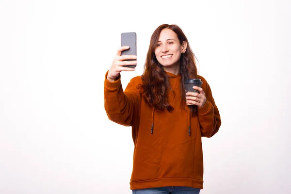 Uma jovem mulher sorrindo está tirando uma foto com um café na mão perto de uma parede branca — Fotografia de Stock
