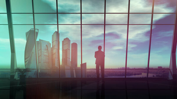 In the office with panoramic windows, a businessman looks at the evening landscape with skyscrapers.
