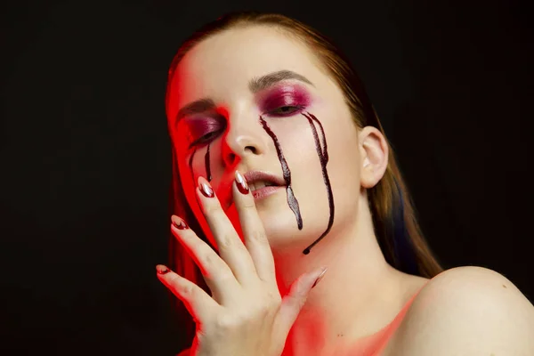 Scary beauty Halloween makeup portrait of a beautiful red hair girl with red eye shadows and fake blood from yellow eyes, studio portrait, red light, black background