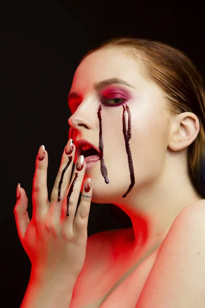 Scary beauty Halloween makeup portrait of a beautiful red hair girl with red eye shadows and fake blood from yellow eyes, studio portrait, red light, black background