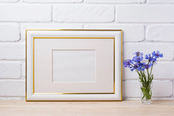 Gold decorated frame mockup with blue cornflower in the simple glass vase. Empty frame mock up for presentation artwork. Template framing for modern art.
