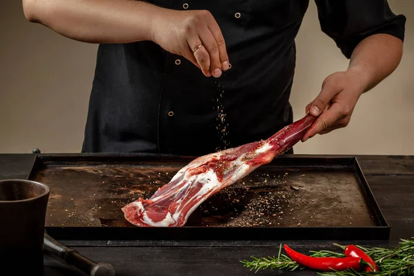 Male chef rubbing raw lamb shanks with salt and pepper on stone tray on wooden table. Chef cooking appetizing shank of lamb. — Stock Photo, Image