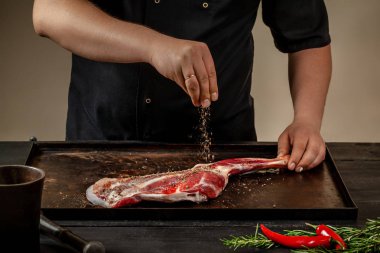 Male chef rubbing raw lamb shanks with salt and pepper on stone tray on wooden table. Chef cooking appetizing shank of lamb. clipart