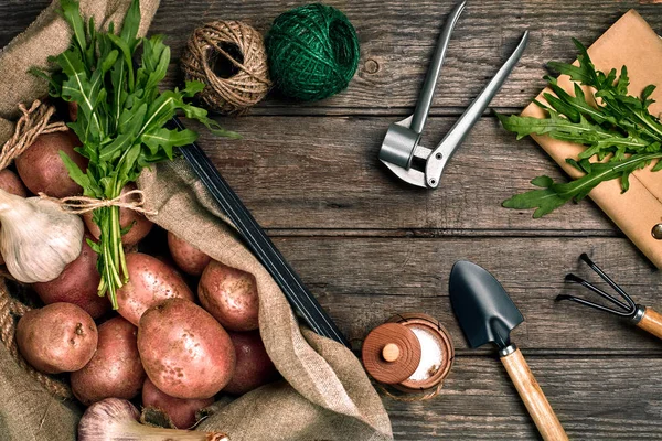 Batatas cruas em um saco de linho, rúcula, alho, pá de jardim e ancinho, fundo de comida, vista superior — Fotografia de Stock