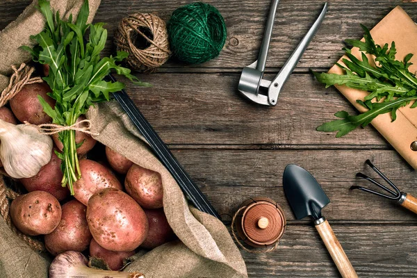 Patatas crudas en una bolsa de lino, rúcula, ajo, pala de jardín y rastrillo, fondo de alimentos, vista superior —  Fotos de Stock