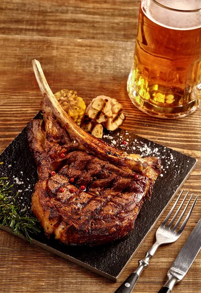 Bife de vaca de cowboy grelhado, copo de cerveja, ervas e especiarias em um fundo de madeira. Vista superior . — Fotografia de Stock