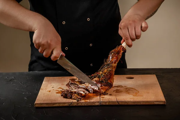 Chef is cutting roasted leg lamb on wooden board on table — Stock Photo, Image