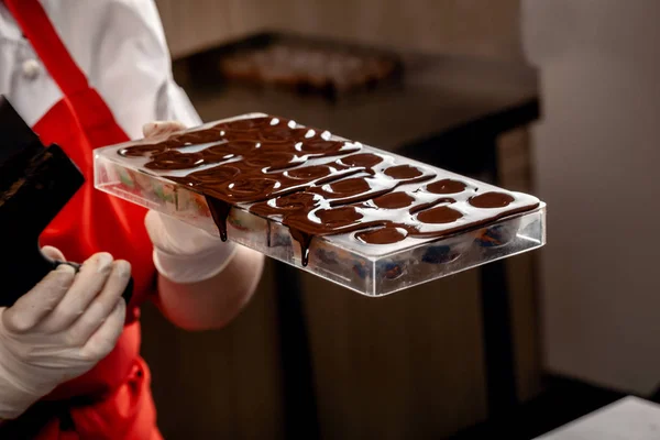 Um confeiteiro de mulher com uniforme vermelho e luvas estéreis brancas faz o grupo de chocolates coloridos do chocolate de leite em uma mesa . — Fotografia de Stock