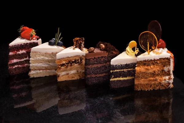 Surtido de grandes trozos de diferentes pasteles: chocolate, frambuesas, fresas, nueces, arándanos. Trozos de pasteles sobre una mesa negra. —  Fotos de Stock