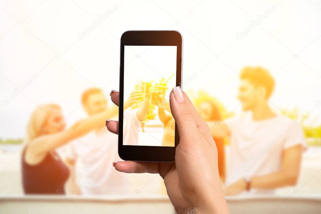 Closely image of female hands holding mobile phone with photo camera mode on the screen. Group of happy friends drinking cocktails at summer bar