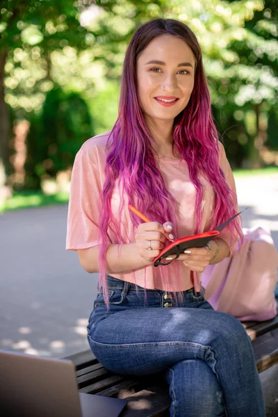 Frau, die in einem Notizbuch auf einer Holzbank im Park sitzt. Mädchen arbeitet im Freien auf tragbarem Computer, Kopierraum. — Stockfoto