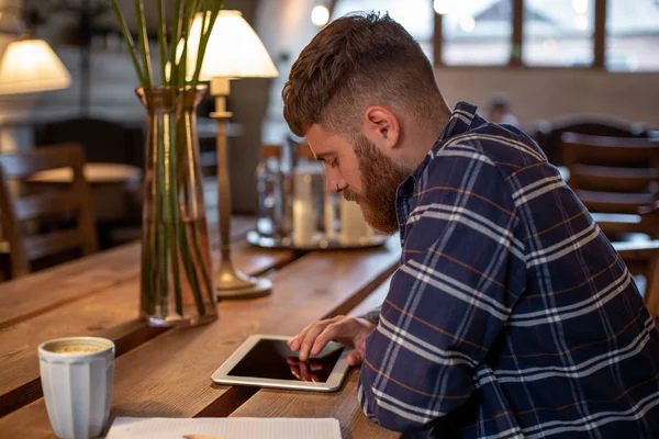 Freelancer vestida con atuendo casual se enfoca en leer noticias y mirar en una tableta digital mientras está sentada en un acogedor café urbano . — Foto de Stock